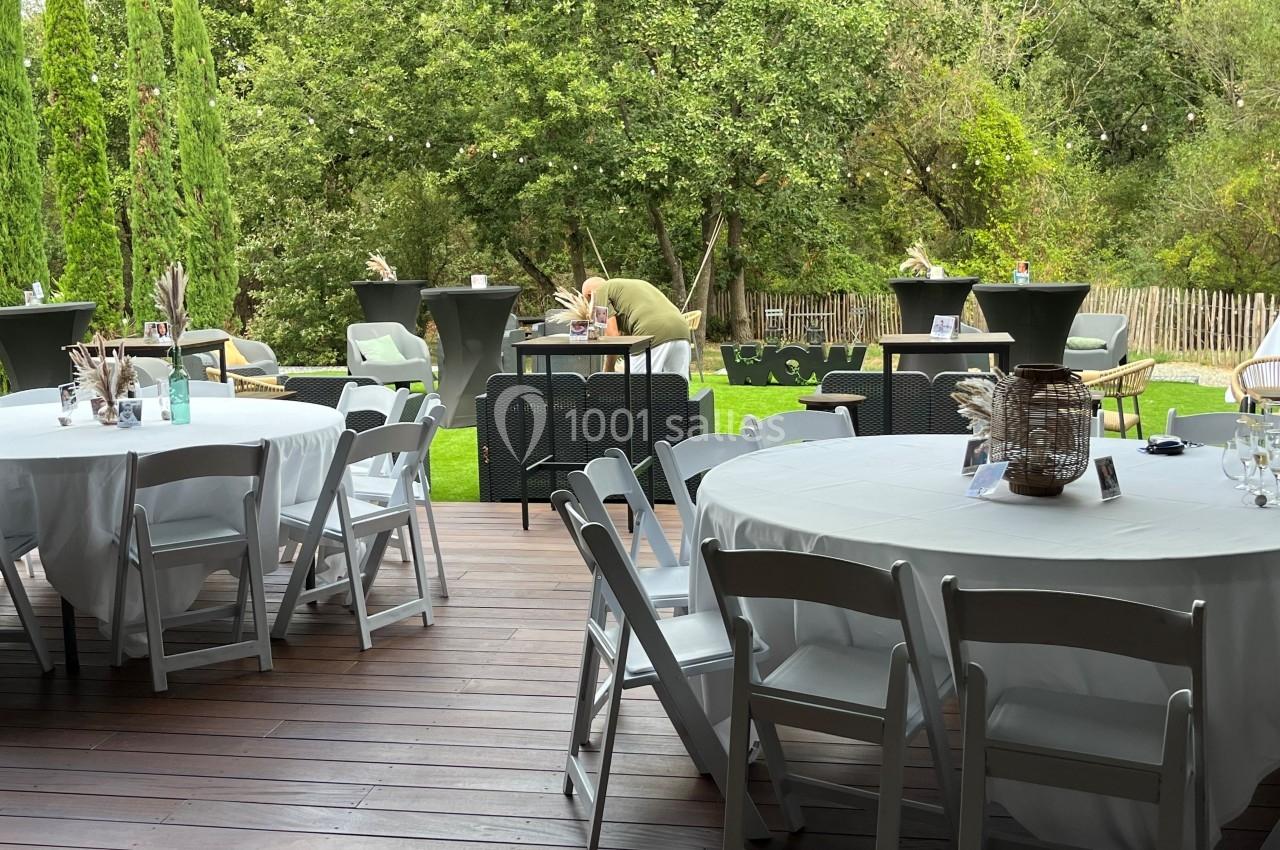 Terrasse en bois avec des tables rondes dressées, chaises blanches et vue sur un jardin verdoyant en arrière-plan.