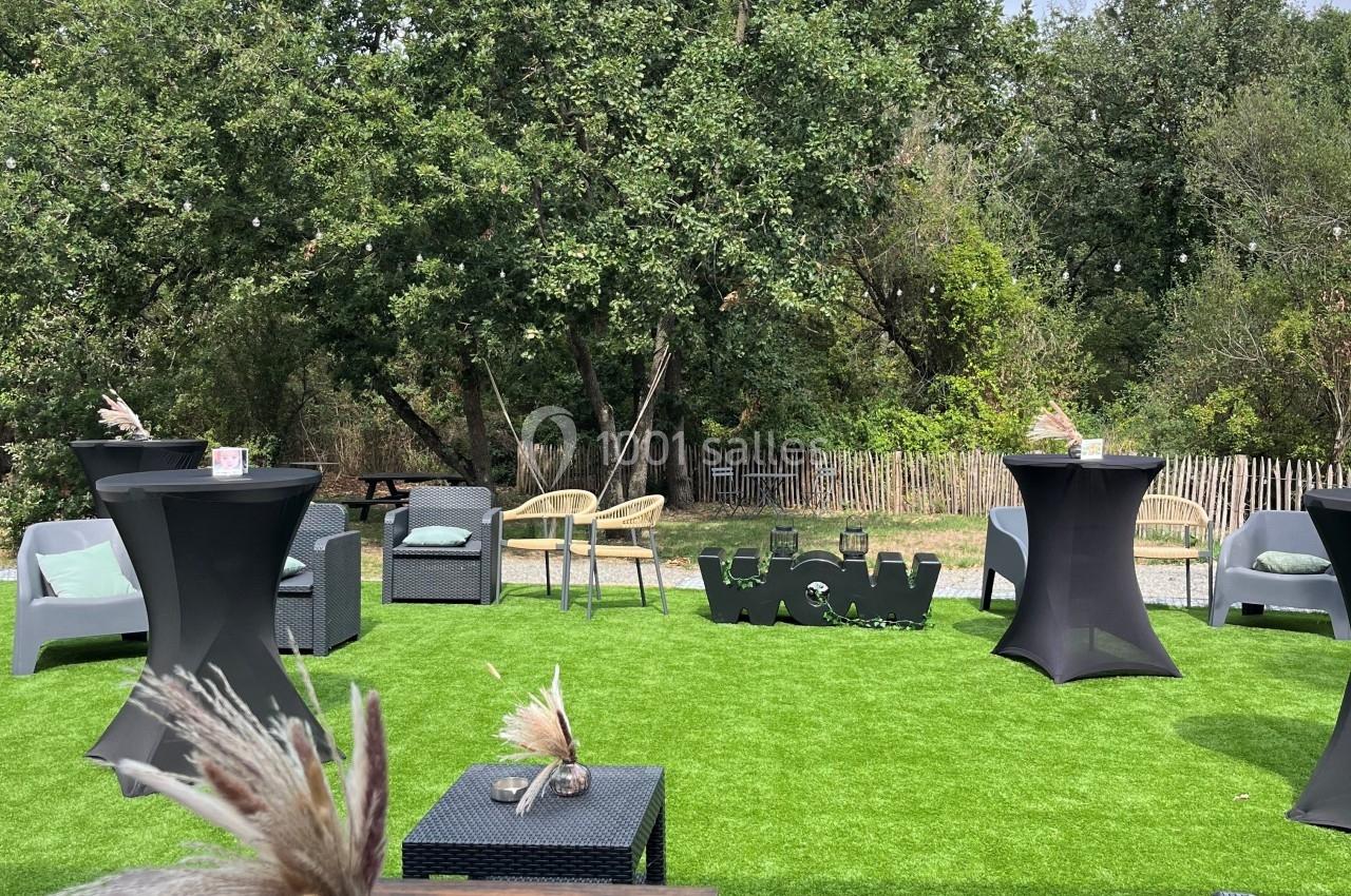 Espace extérieur avec tables, fauteuils et mange-debout sur pelouse artificielle, entouré d'arbres et de végétation.