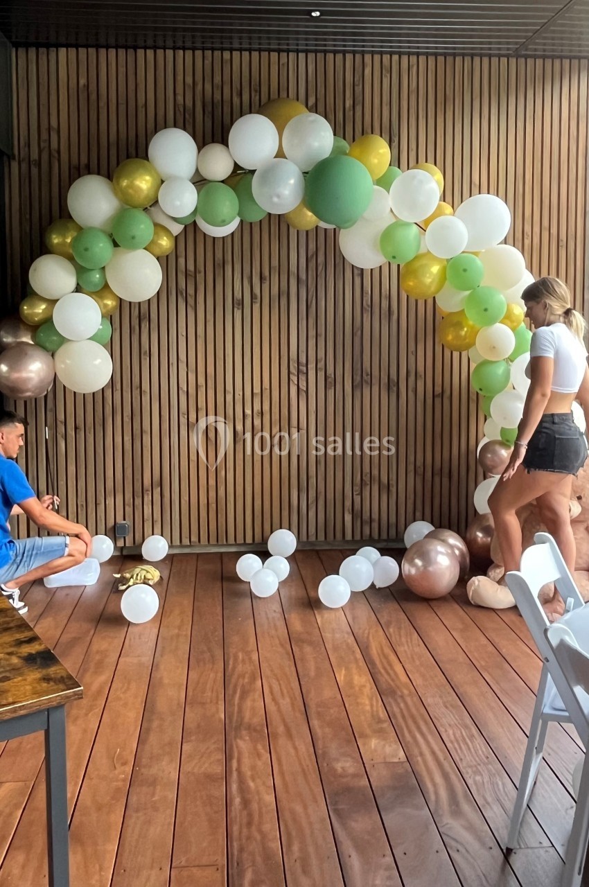 Deux personnes assemblent une arche de ballons colorés sur une terrasse en bois, près d'un mur en lambris.