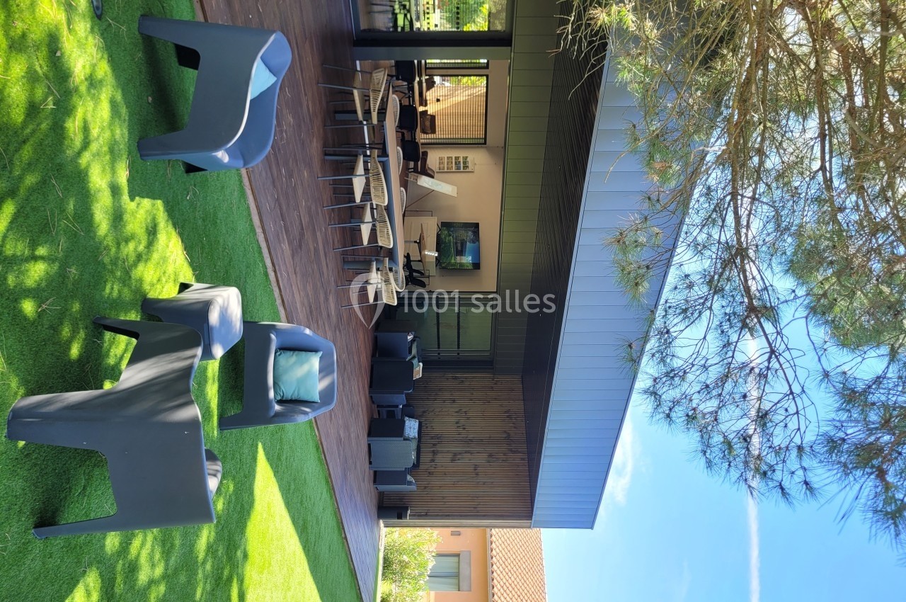 Terrasse moderne avec mobilier en plastique et bois, entourée de verdure et partiellement ombragée par un arbre.