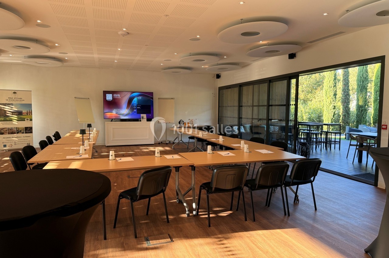 Salle de réunion lumineuse avec tables disposées en U, écran de présentation et vue sur une terrasse extérieure arborée.