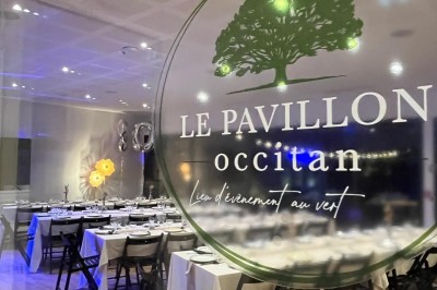 Chambre lumineuse avec lit double, bureau, coin salon, kitchenette et vue sur un jardin à travers une baie vitrée.