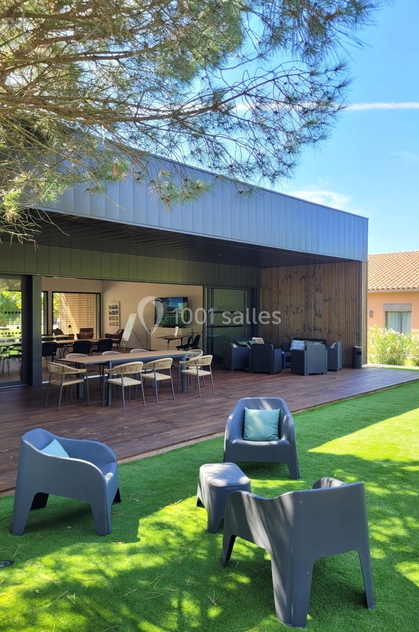 Terrasse moderne avec mobilier de jardin, espace repas et salon, entourée de pelouse et d'arbres sous un ciel dégagé.