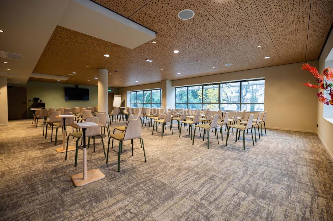 Salle de réunion lumineuse avec des chaises disposées en rangées, grandes fenêtres et plafond en bois perforé.
