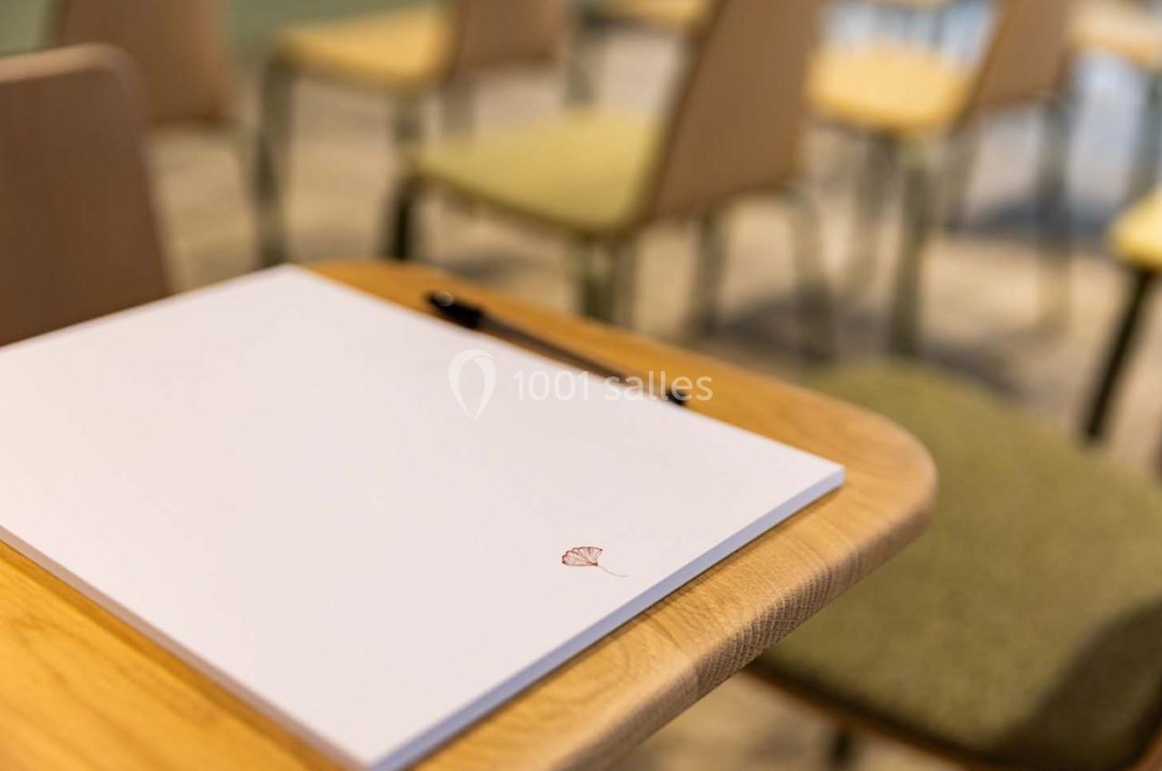 Bloc-notes blanc avec un stylo noir posé sur une table en bois, entouré de chaises floues en arrière-plan.