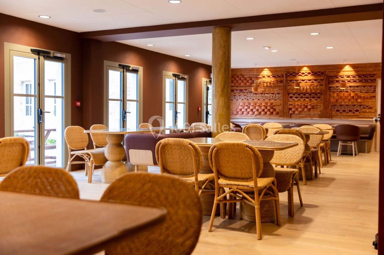 Salle lumineuse avec tables et chaises en rotin, décorée de boiseries murales et baignée de lumière naturelle.