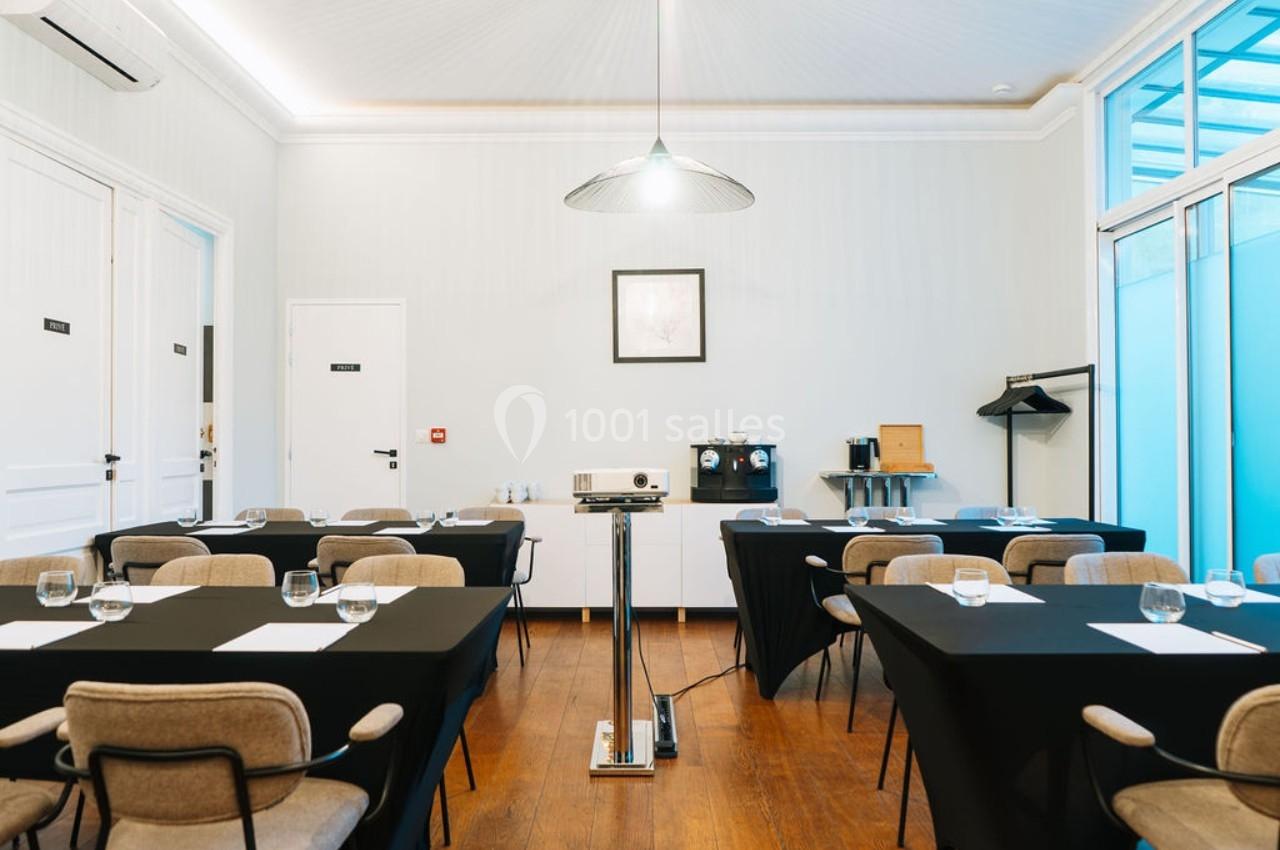 Salle de réunion lumineuse avec tables noires, chaises, projecteur central et coin café en arrière-plan.