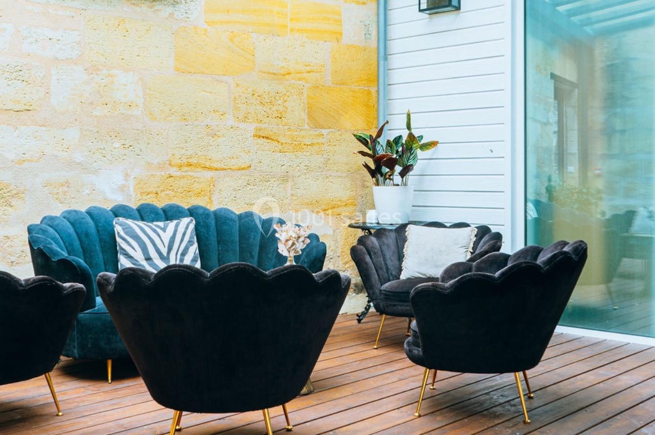 Salon de jardin avec fauteuils noirs, coussins décoratifs et plantes, installé sur une terrasse en bois près d'un mur en…