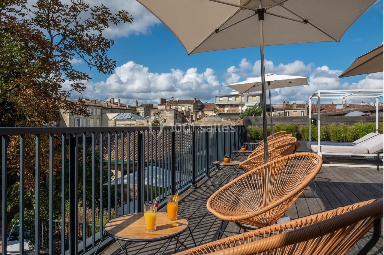 Terrasse ensoleillée avec fauteuils en rotin, tables basses et parasols, offrant une vue sur les toits d'une ville.