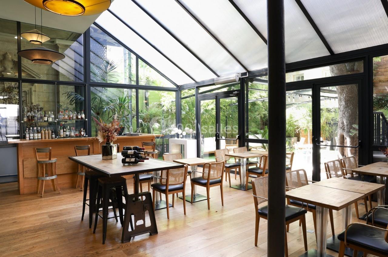 Salle lumineuse avec verrière, mobilier en bois, bar en arrière-plan et vue sur un jardin verdoyant.