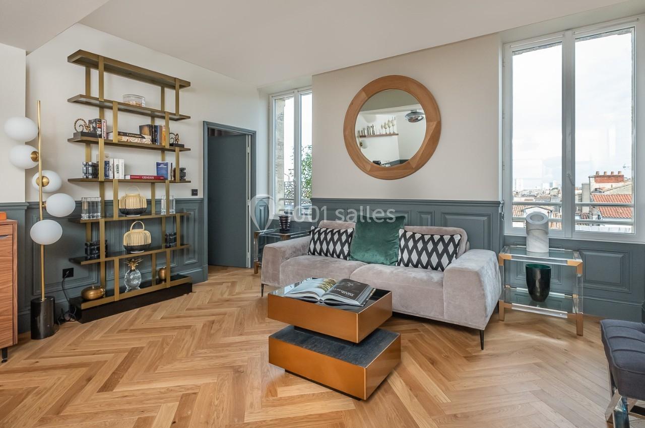 Salon lumineux avec parquet en chevrons, canapé beige, étagères modernes, miroir rond et grandes fenêtres.