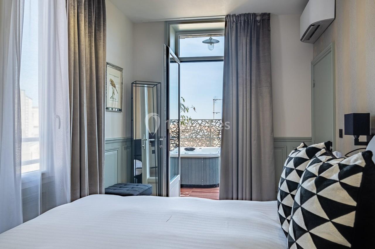 Chambre lumineuse avec lit double, rideaux gris, miroir, balcon ouvert et vue sur un jacuzzi extérieur.