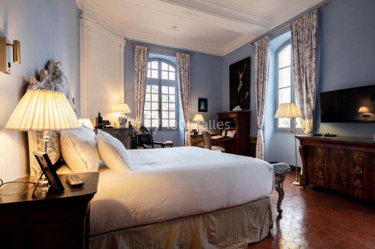 Chambre élégante avec lit double, meubles anciens, lampes allumées et grandes fenêtres laissant entrer la lumière naturelle.