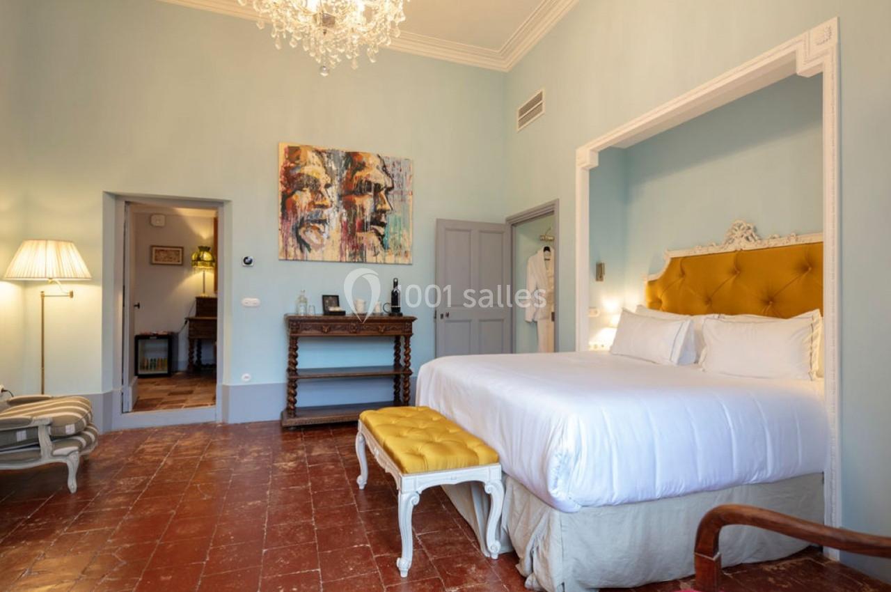 Chambre lumineuse avec lit double, tête de lit jaune, banc assorti, lustre, tableau coloré et sol en terre cuite.