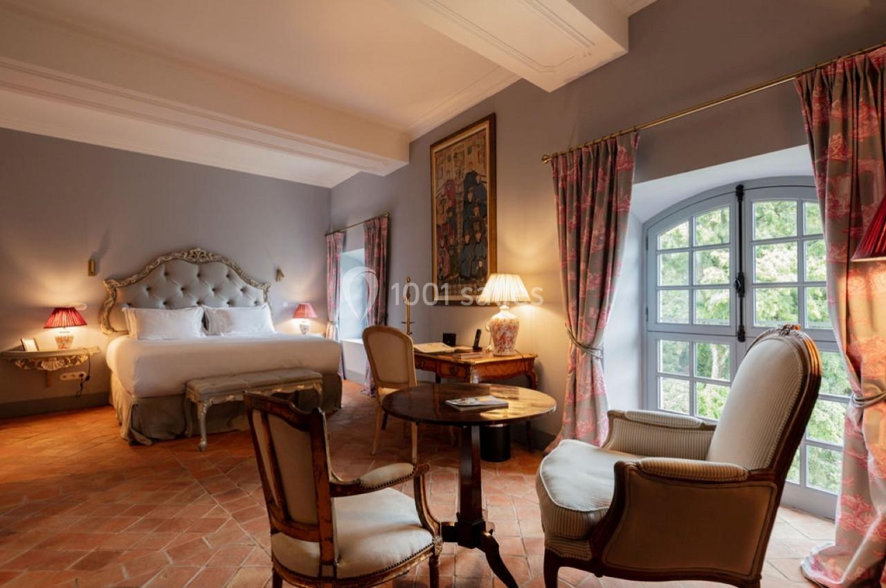 Chambre élégante avec lit double, fauteuils, table en bois, grandes fenêtres et lumière naturelle.