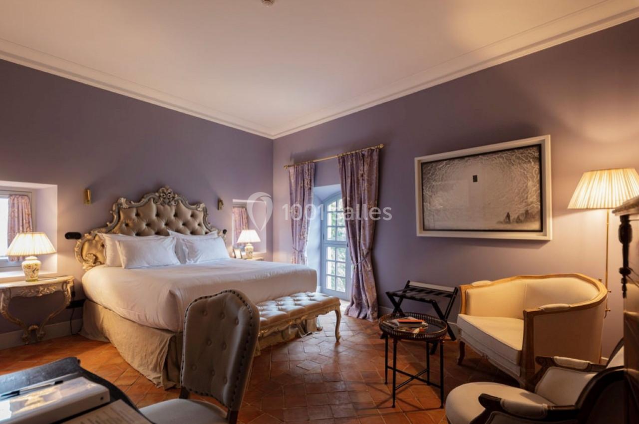 Chambre élégante avec lit double, mobilier classique, murs lavande, éclairage doux et fenêtres avec rideaux.