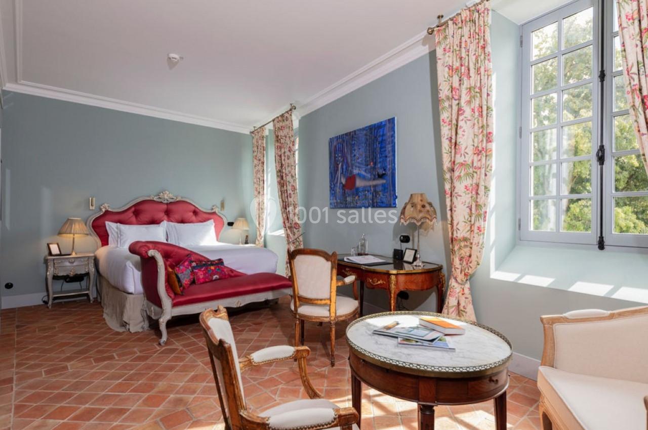 Chambre lumineuse avec lit double, fauteuils, table ronde, bureau, sol en terre cuite et fenêtre donnant sur un jardin.