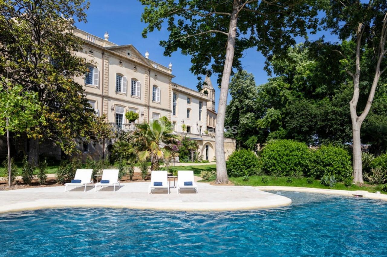 Façade d'un bâtiment historique entouré de verdure, avec une piscine et des transats au premier plan.