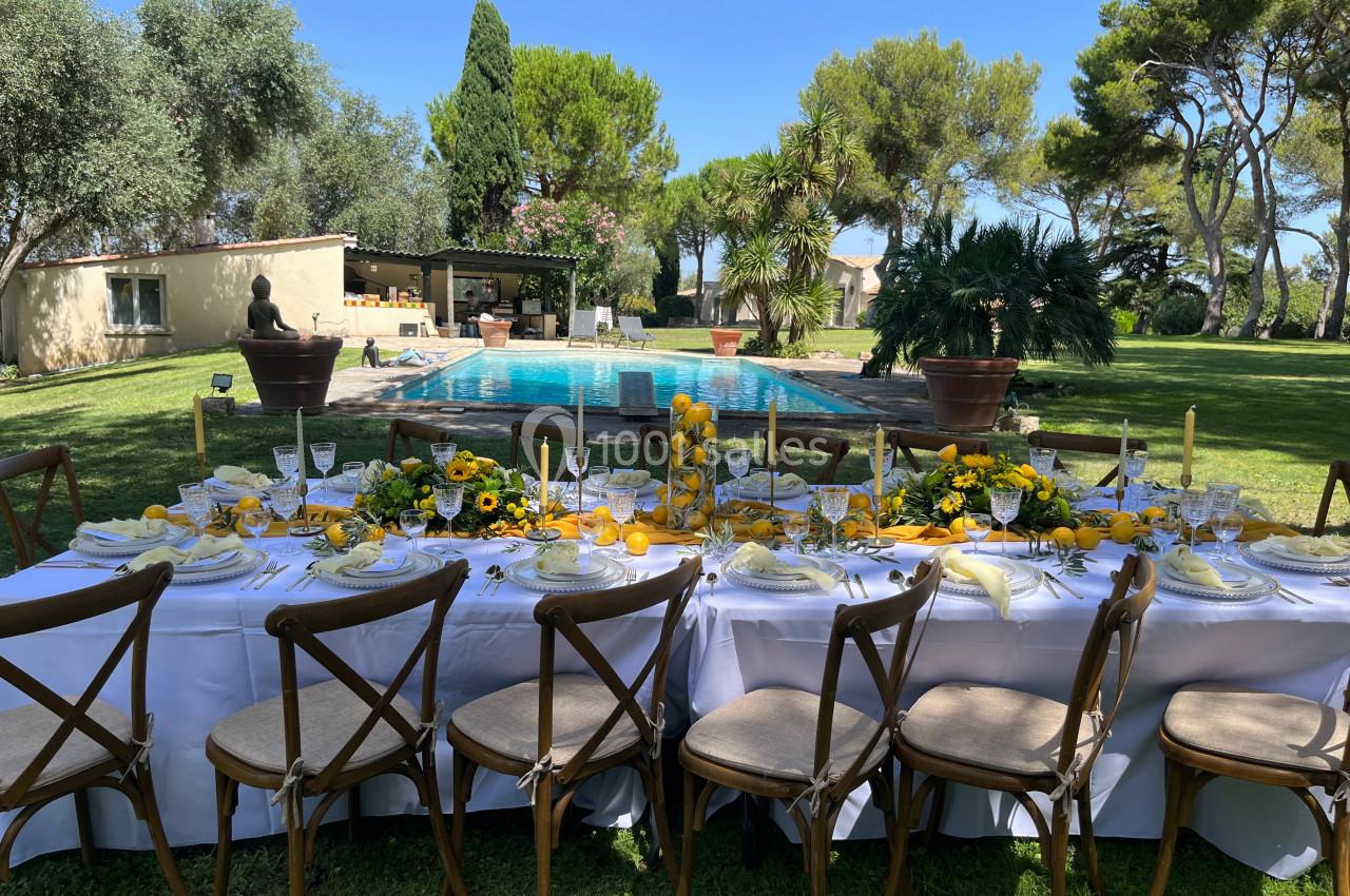 Table dressée en extérieur avec une décoration florale jaune, face à une piscine entourée de verdure et d'arbres.