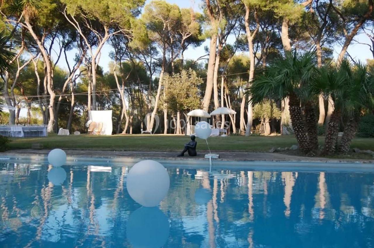Piscine entourée d'arbres et de palmiers, décorée de ballons blancs flottants et de parasols en arrière-plan.