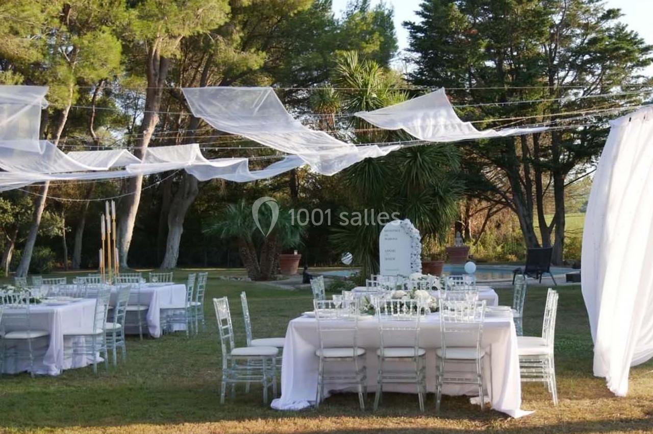 Tables décorées pour un événement en plein air, entourées de verdure et recouvertes de voiles blancs suspendus.