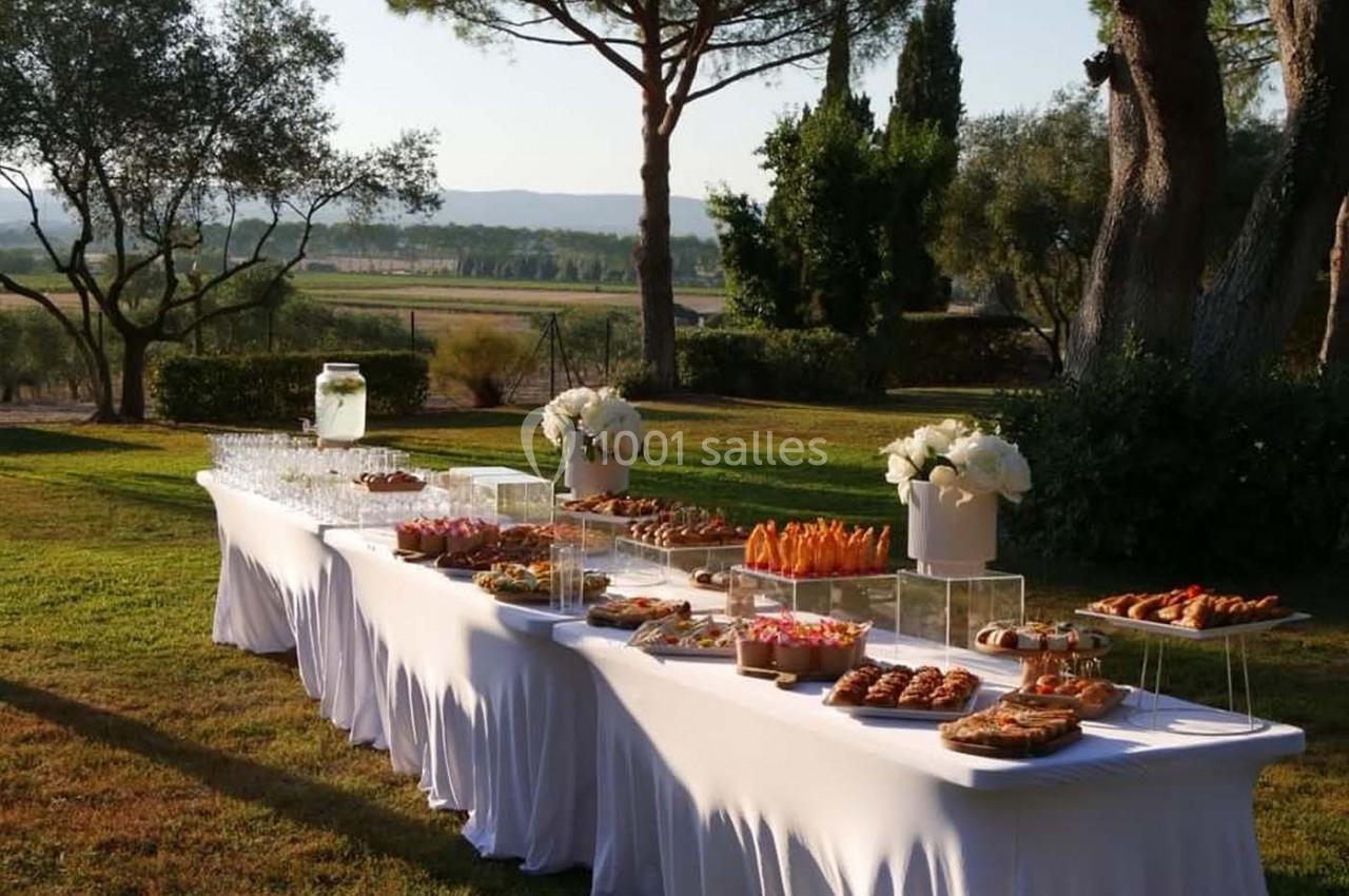Table dressée en extérieur avec nappes blanches, garnie de viennoiseries, desserts et boissons, entourée de verdure.
