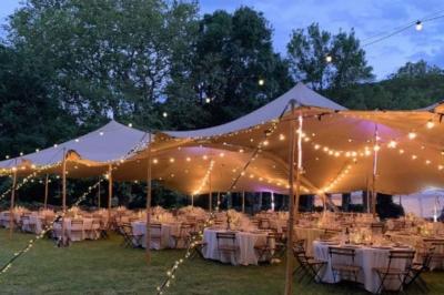 Grand chapiteau beige éclairé par des guirlandes lumineuses, abritant des tables rondes dressées pour un événement en plein…