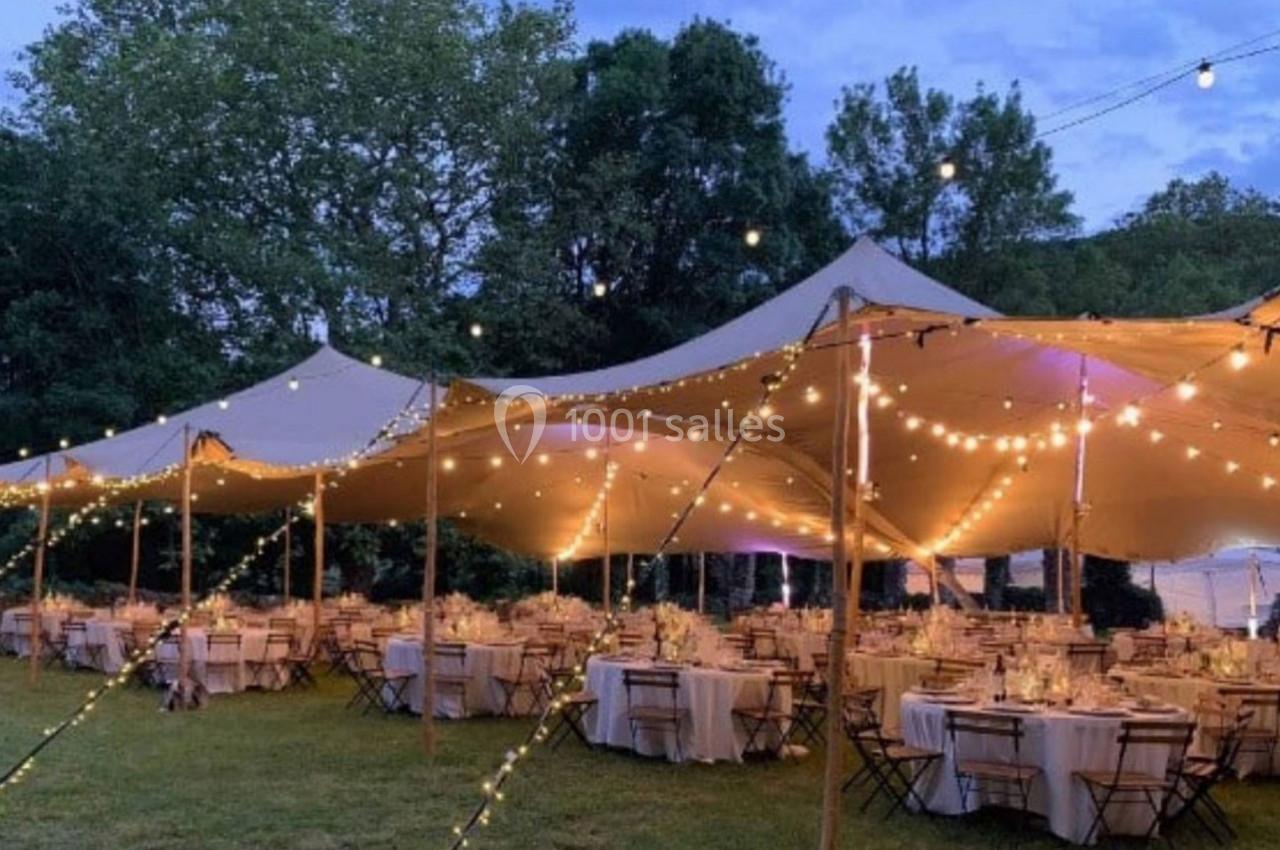 Grand chapiteau beige éclairé par des guirlandes lumineuses, abritant des tables rondes dressées pour un événement en plein…