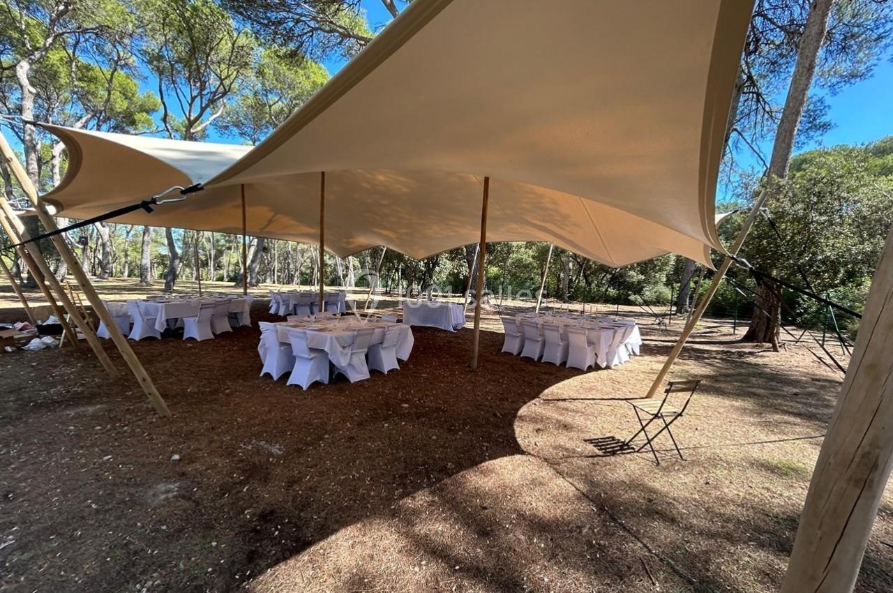 Tente beige montée en forêt, abritant des tables et chaises blanches disposées pour un événement en plein air.
