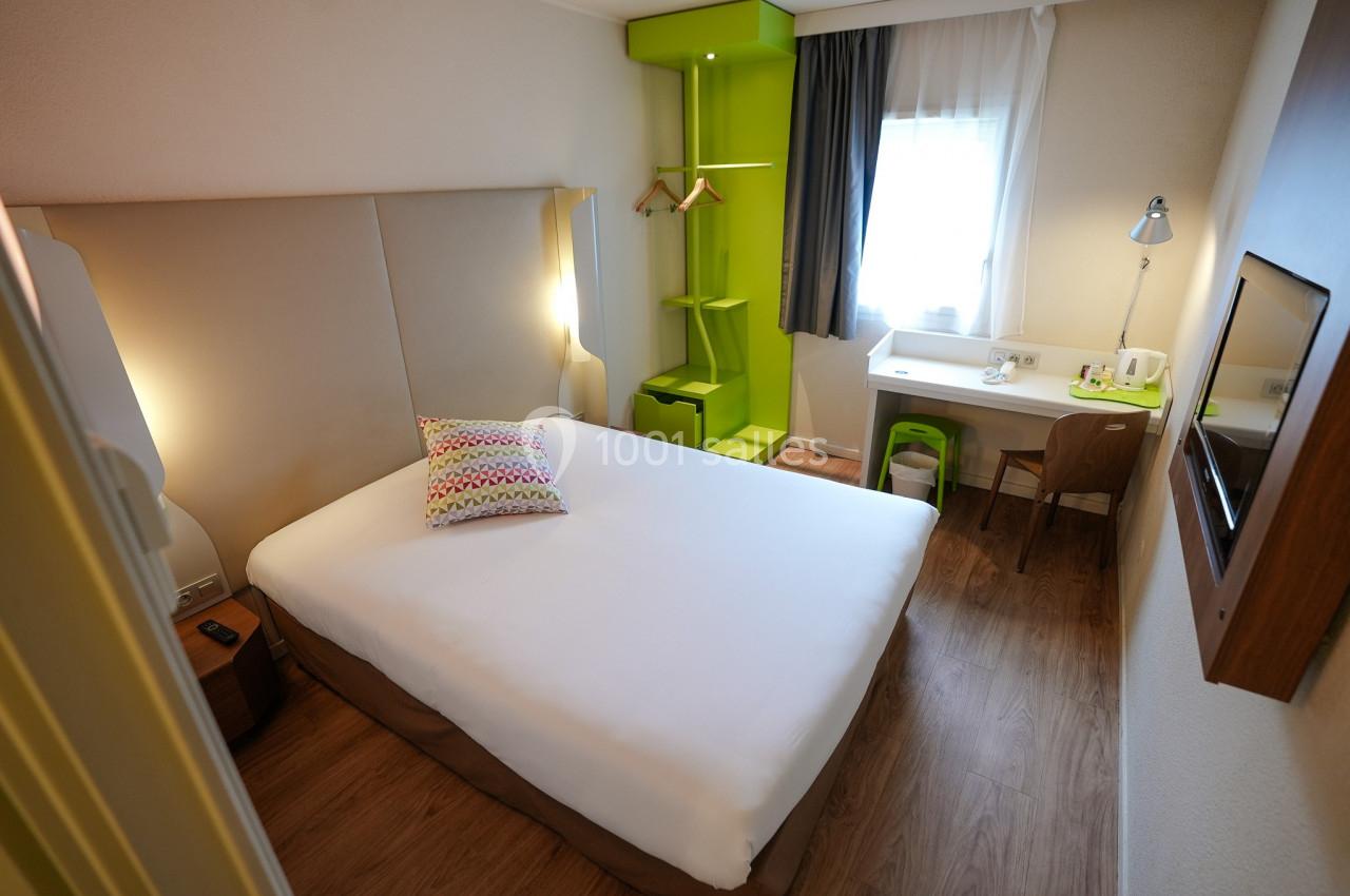 Chambre d'hôtel lumineuse avec lit double, bureau, chaise, étagères vertes et fenêtre avec rideaux.