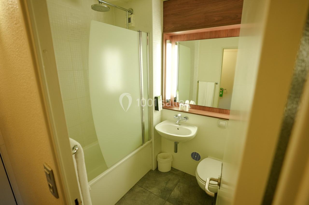 Salle de bain avec baignoire-douche, lavabo, miroir éclairé, toilettes et carrelage au sol.