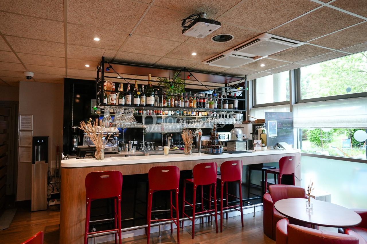 Bar moderne avec comptoir en marbre, tabourets rouges, étagères garnies de bouteilles et grande fenêtre lumineuse.