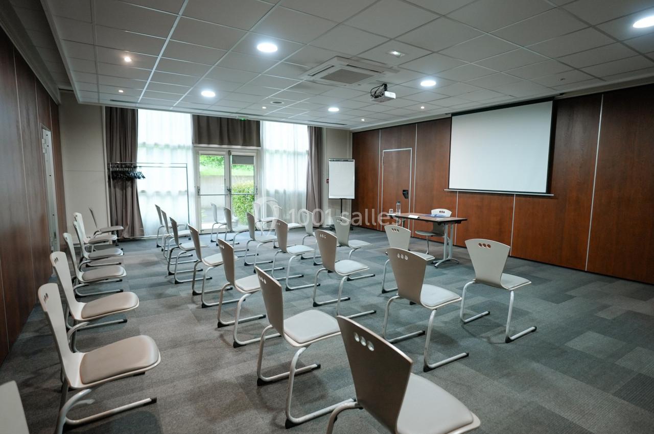 Salle de réunion lumineuse avec des chaises alignées, un projecteur, un écran et un tableau blanc.