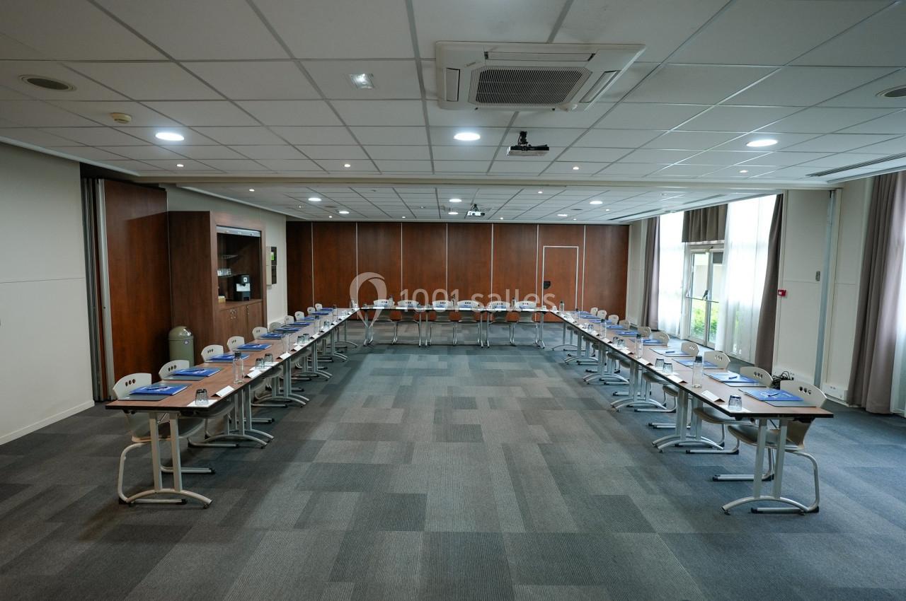 Salle de réunion spacieuse avec disposition en U, tables équipées de blocs-notes et chaises, éclairée par des fenêtres.