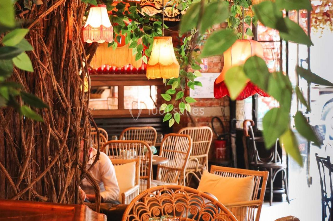 Intérieur d'un café chaleureux avec des fauteuils en rotin, des lampes suspendues et une décoration végétale abondante.