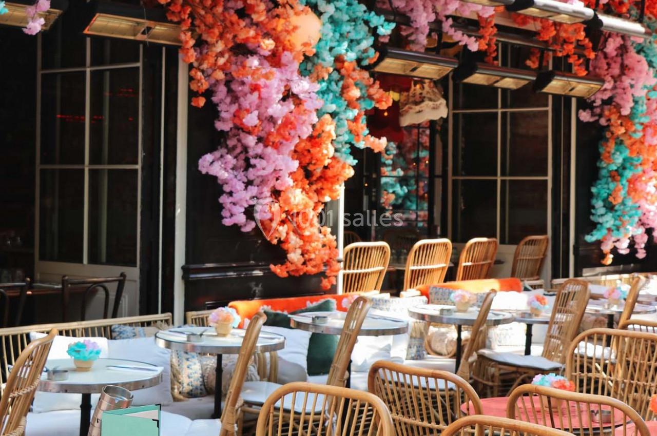 Terrasse de café décorée de fleurs colorées avec des chaises en rotin et des tables dressées en plein air.