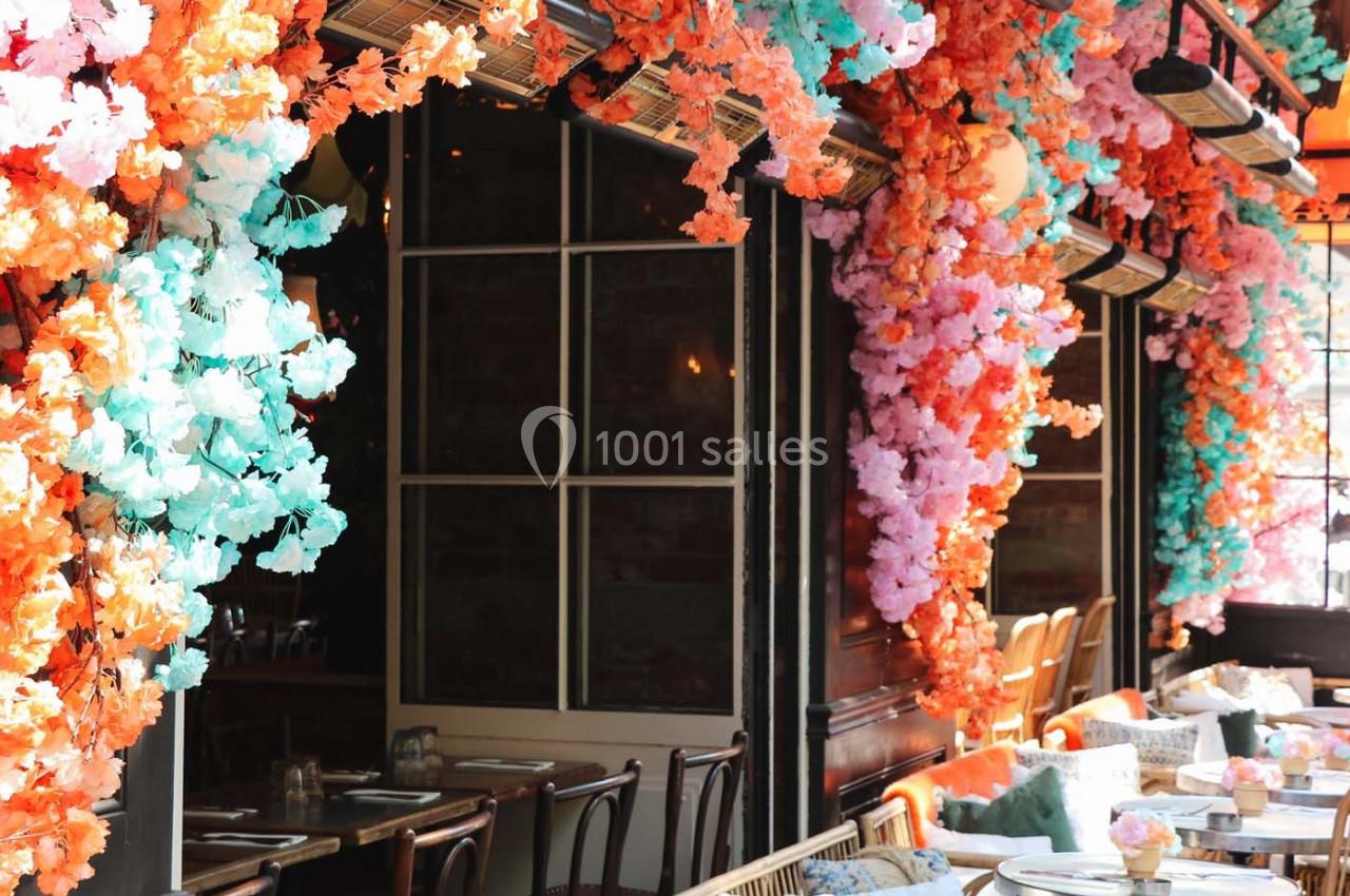 Façade de restaurant décorée de fleurs artificielles colorées avec des tables dressées en terrasse.