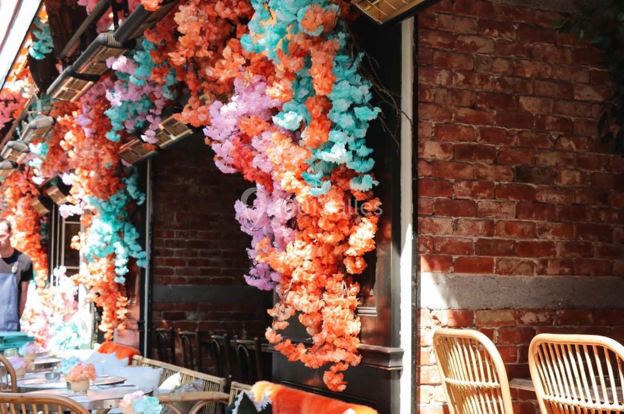 Guirlandes de fleurs colorées suspendues à une façade en briques, au-dessus de tables et chaises en terrasse.