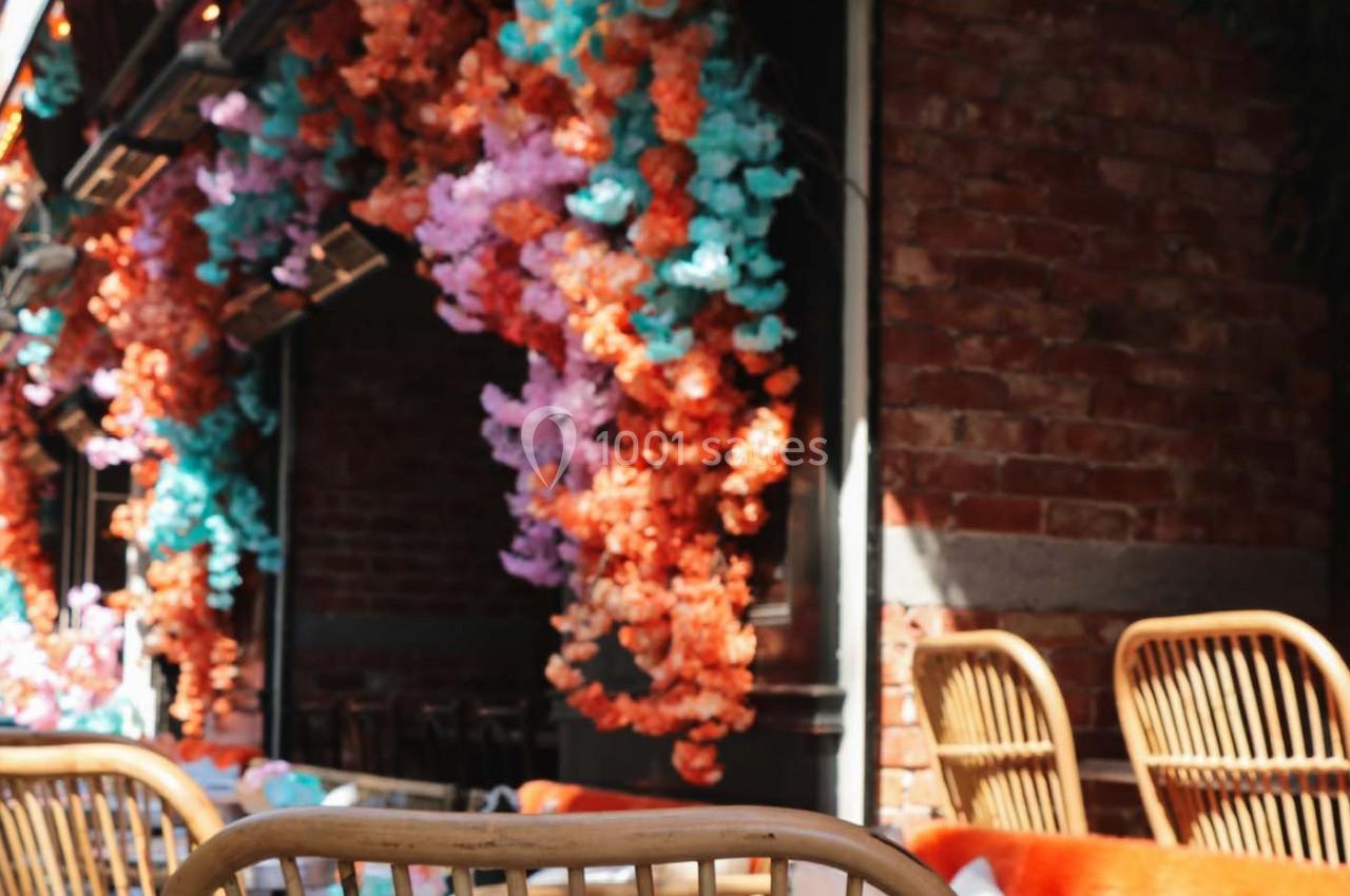 Façade en briques ornée de guirlandes de fleurs colorées avec des chaises en rotin au premier plan.