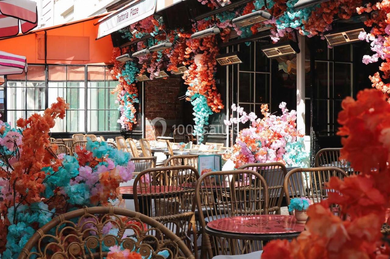 Terrasse de café décorée de fleurs colorées, avec des tables en rotin et une lumière naturelle abondante.