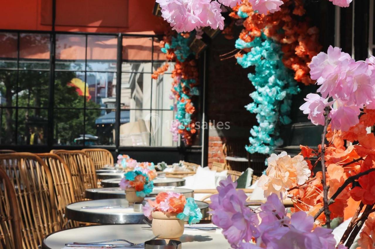 Terrasse décorée de fleurs colorées avec des tables rondes et des chaises en rotin, baignée de lumière naturelle.