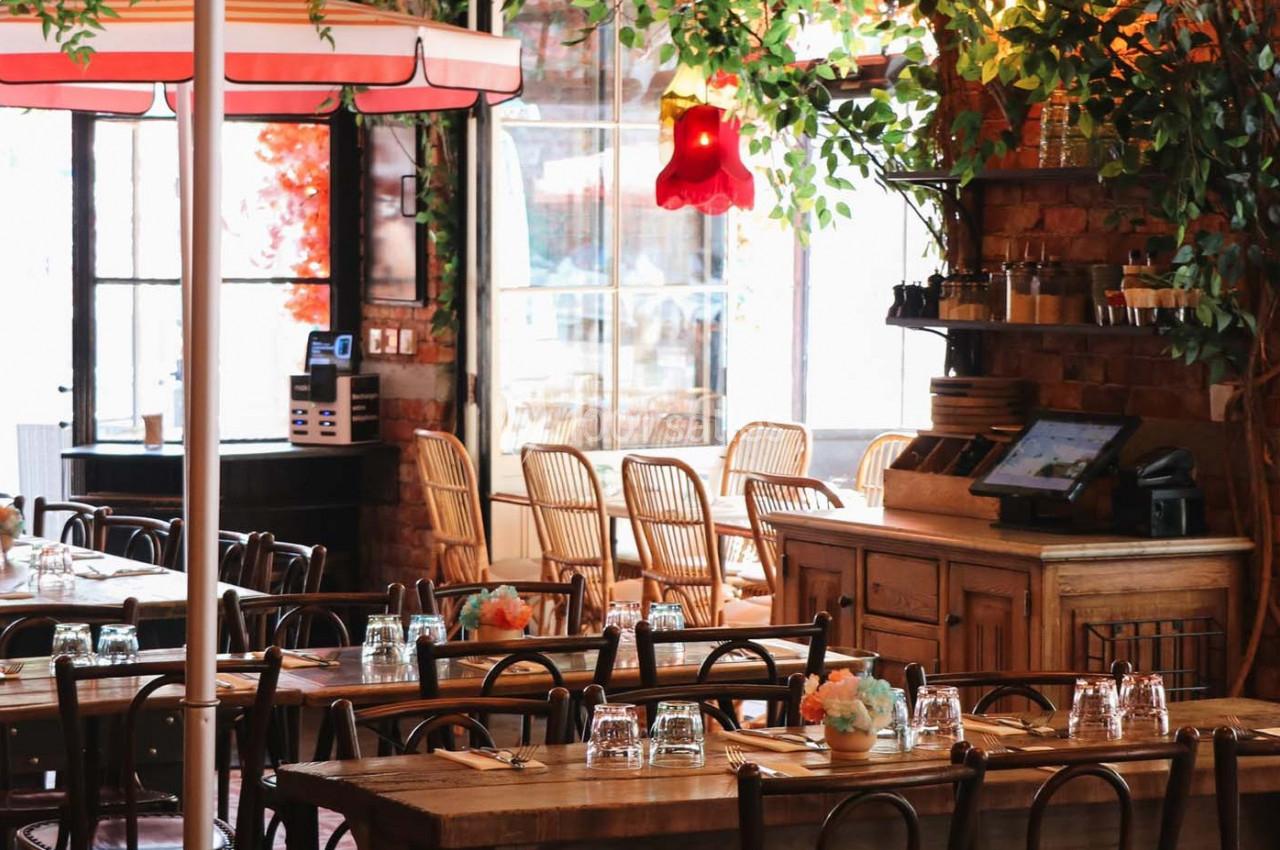 Salle de restaurant lumineuse avec tables en bois, chaises en rotin, plantes décoratives et une caisse enregistreuse.