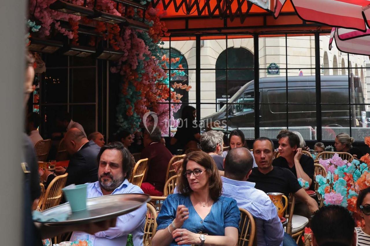 Des personnes assises à des tables en terrasse, entourées de décorations florales colorées, dans un cadre urbain.