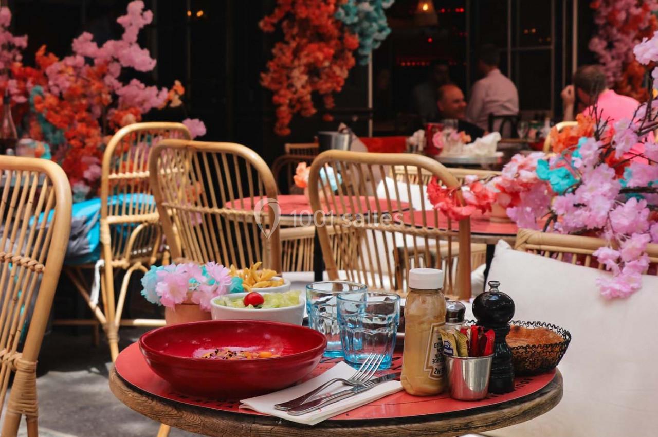 Table en terrasse avec assiette de nourriture, couverts, verres et décor floral coloré en arrière-plan.