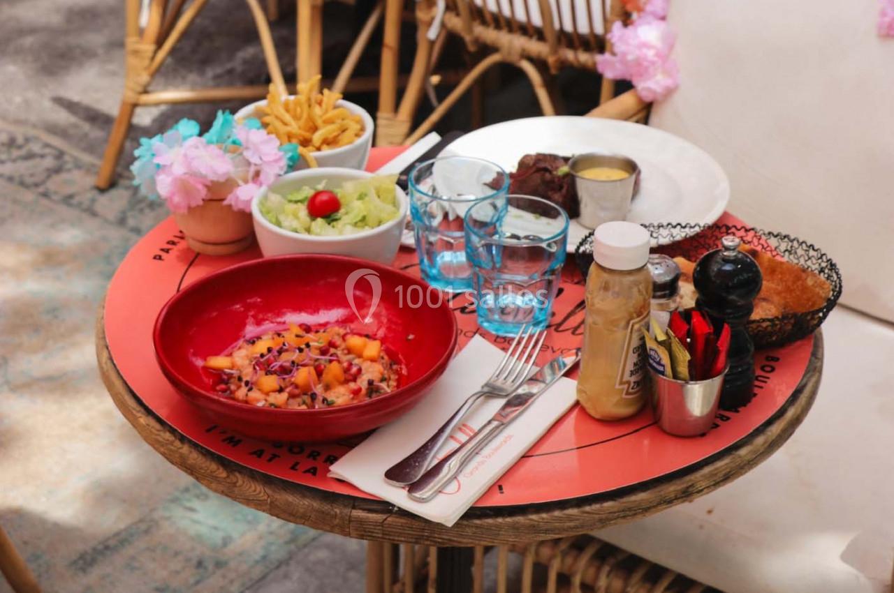 Table ronde avec un repas composé de salade, viande, frites, sauces et boissons, servie en extérieur sur une terrasse.