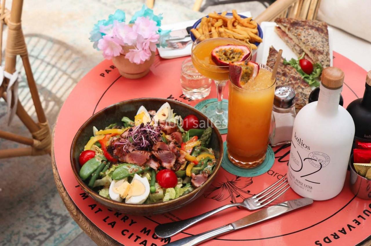 Assiette de salade composée avec légumes, œufs et viande, entourée de boissons, frites et pain sur une table colorée.
