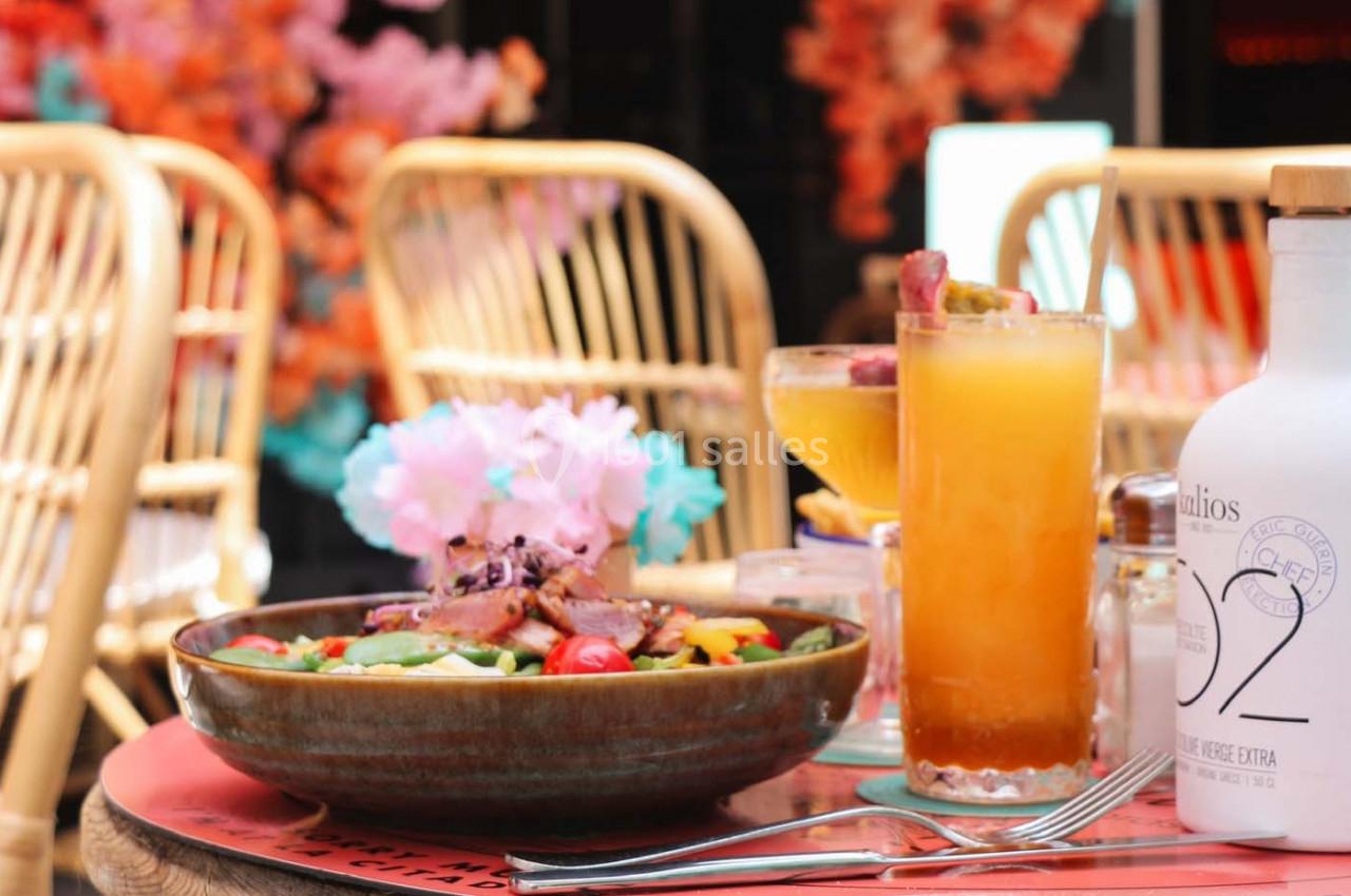 Assiette de salade colorée accompagnée de cocktails et d'une bouteille d'huile sur une table en terrasse.