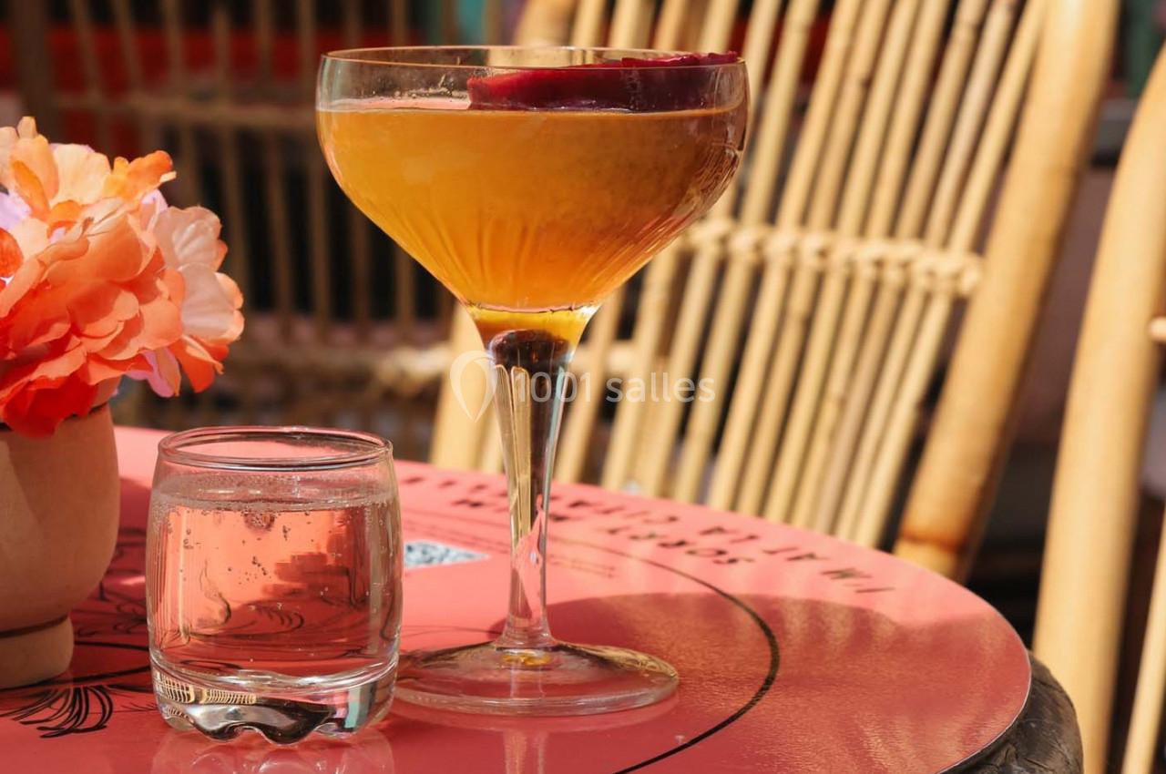 Verre à cocktail orange avec une tranche de fruit, posé sur une table rose près d'un verre d'eau et de fleurs.