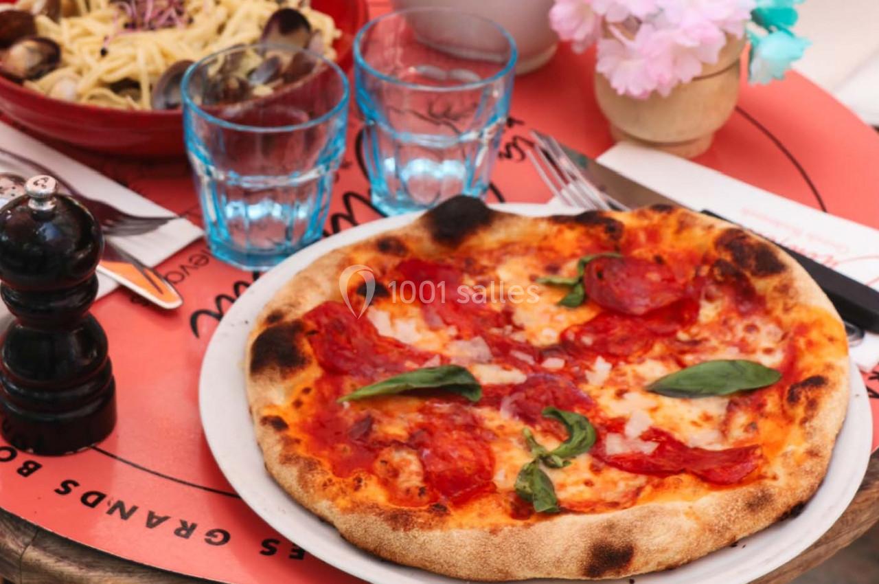 Pizza garnie de tranches de chorizo, basilic et fromage, servie sur une table avec des verres et des couverts.