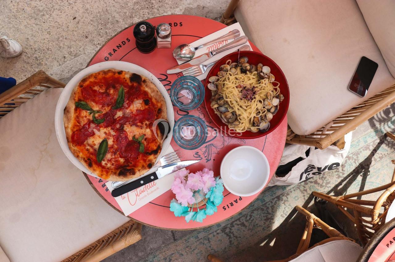 Table ronde avec une pizza garnie de basilic, un plat de pâtes aux palourdes, des verres et des couverts.